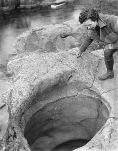 A woman identified as Beth Doane is inspecting a large pothole in a rock formation high above the St. Croix River in Interstate Park. She is wearing gloves, a heavy jacket and knee-high lace-up boots. There is a boat on the river in the background.  