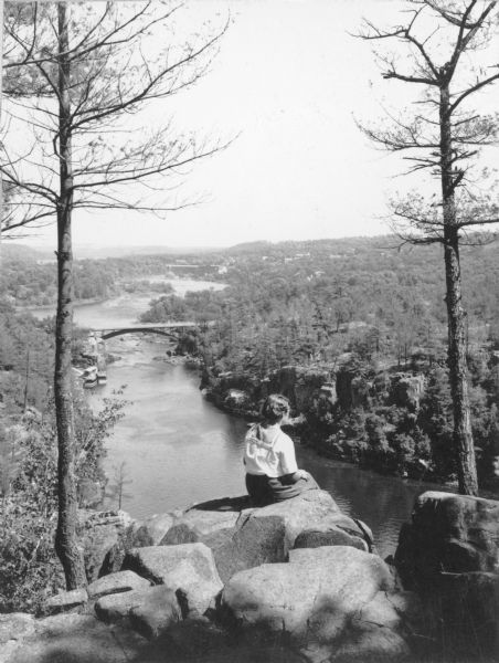 View towards an unidentified woman sitting and facing away from the camera to take in the view of the St. Croix River from a high rock formation. The bridge between Wisconsin and Minnesota, located at the northern end of the Dalles of the St. Croix, is in the middle background. In the distance is the St. Croix Falls hydroelectric dam and power plant, built 1905-1907.  