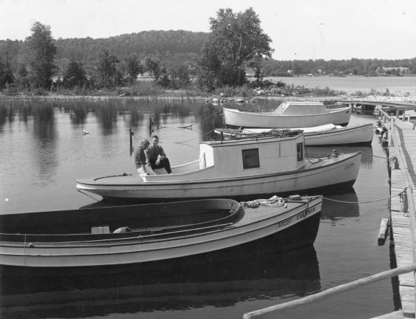 View from pier towards a young couple sitting in the back of a wooden boat tied to a pier in Peninsula State Park. Fish Creek Harbor is in the background, and the village of Fish Creek is obscured by trees. Three other boats are moored to the pier. 