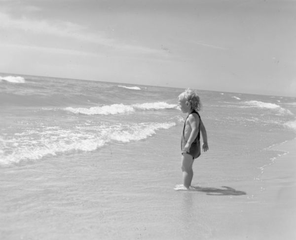 A child wearing a bathing suit is standing with their feet in shallow water as waves approach the shore. On the reverse of the print is written: "A tiny tot surveys the expanse of Lake Michigan from the sandy beach at Terry Andrae State Park, Wisconsin."