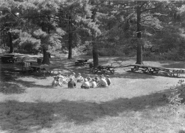 Fourteen Boy Scouts are sitting in a circle in a clearing at Terry Andrae State Park. One boy on the right is older and is shirtless. There are picnic tables and mature pine trees in the background.