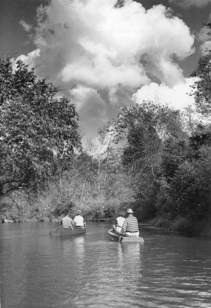 View across water towards two men in a canoe on the left, and a man and woman in a duckboat on the right paddling down the Clam River. Trees and shrubs cover the riverbank. There are large cumulus clouds overhead.