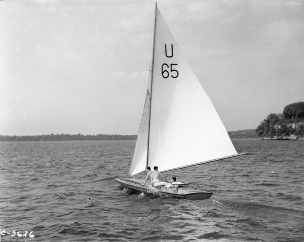 Rear view of three young men sailing an E-Scow sailboat numbered U 65 in a regatta on Lake Mendota. "NAN TUCK" is painted on the stern of the boat. Maple Bluff is in the background.