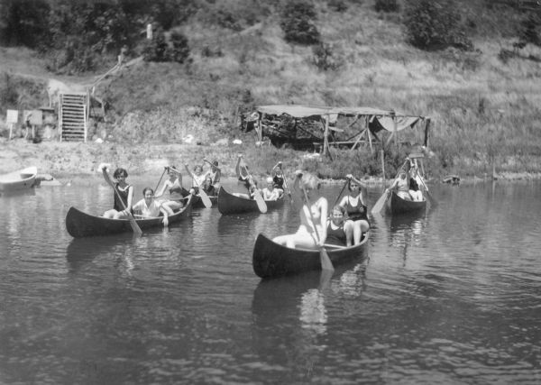 View across water towards Girl Scouts in five canoes setting out from shore on Rice Lake at Camp Hickory Hill. There is a temporary shade structure and wooden steps at the base of a hill in the background. Most of the scouts are wearing bathing suits.
