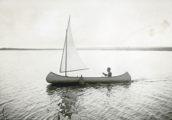 A young man is sitting and holding the mainsheet in a canoe fitted with a single mast and sail. There is a small wake behind the boat. The far shoreline is in the distance.
