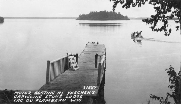 A motor boat carrying three people is leaving a small wake in the calm waters of Crawling Stone Lake as it heads toward an island. There is a worn wooden pier in the forground. The caption reads: "Motor boating at Yeschek's Crawling Stone Lodge Lac du Flambeau Wis."