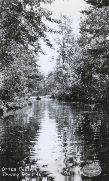 A picturesque view of three people in a rowboat on Otter Creek, with Government Island in the background. A seal at lower right identifies Estberg Photo Service and "Chain o' Lakes Waupaca, Wis. The Killarneys of America."   