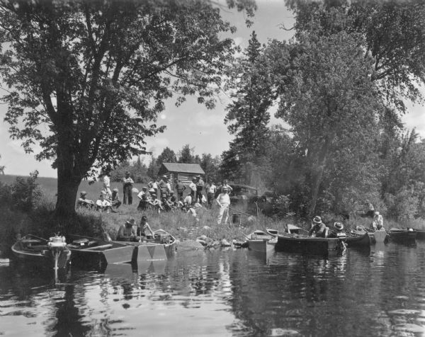 View across water towards a shoreline where men and boys are gathered on the bank of the Flambeau River, with several canoes and rowboats pulled up to the rocky shore. Some of the boats are fitted with outboard motors. A large covered kettle is suspended from a tripod over a campfire. There is an automobile and a small log cabin in the background. A caption on the reverse of the print reads: "River trip on Flambeau River from Turtle Flowage to Park Falls, sponsored by Flambeau Rod & Gun Club." 