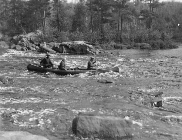 View across river towards three men in a canoe negotiating the Cedar Rapids of the Flambeau. Large boulders are along the far shoreline. On the reverse of the print is written: "Warden [Allen] Hanson, Commissioners [L.M.] Hobbins and [Edwin Monroe] Dahlberg, shooting the rapids." 
