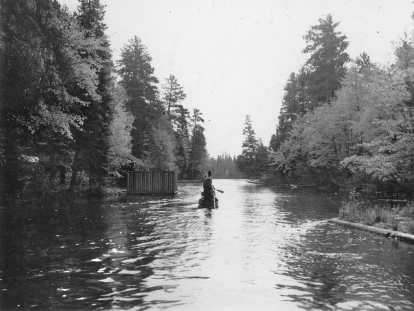Rear view of two canoes, one directly behind the other, each propelled by a man traveling on the Brule River. The man in the foreground is using a pole, and in the far canoe the man is using a paddle. There is a structure with vertical log walls on a pier on the left; the roof of a cabin is visible beyond the structure.