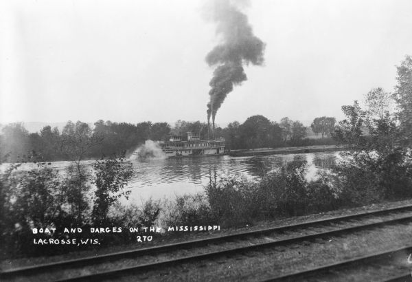 View across railroad tracks along the shorline of the Mississippi River towards the stern wheeled steamboat <i>General Allen</i> towing barges upriver at LaCrosse. Dark smoke is pouring from the two stacks. The initials "U.S." are painted on the wheelhouse.