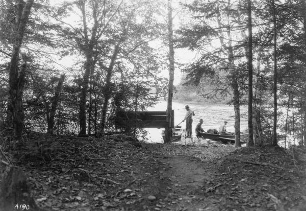 View from among trees towards three men working to pull up and secure three rowboats at a rustic log landing site on the Flambeau River. One man is still sitting in one of the boats which is also holding two packs and what appears to be a suitcase. There is a wooden sign attached to trees at the river's edge.