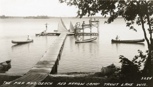 View from shoreline towards a long pier on Trout Lake leading to a diving board on the left, and diving platforms on the right. There are small sailboats tied up at the pier, and three canoes are pulled up on the shoreline. Three people are each paddling their own canoe, a group of boys are sitting on the platforms, and one boy is standing on the diving board.