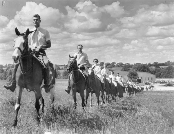View towards a line of several dozen men and women, in equestrian attire and on horseback, riding single file up a hill from a large arena style building set in rolling terrain. There are cars in a parking lot adjacent to the building. On the reverse of the print is written: "Good riding horses are plentiful in Wisconsin."