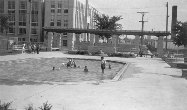 A view, looking south, at the intersection of West Mt. Vernon Avenue and North 65th Street. In the foreground is a shallow pool where children are playing, some wearing street clothes and others in bathing suits and caps. Other children are standing on the pavement at left; there are older girls or young women at right. Beyond the pool is a tiered fountain set into the wall of a walkway shaded by a pergola. On the far left is a sign which reads: "USA WORK PROGRAM WPA." The large brick building at left is Solomon Juneau High School. On the right a tall smokestack is towering over the greenhouses of Kennedy and Kennedy Florists. The houses in the far background are on West Fairview Avenue.  
