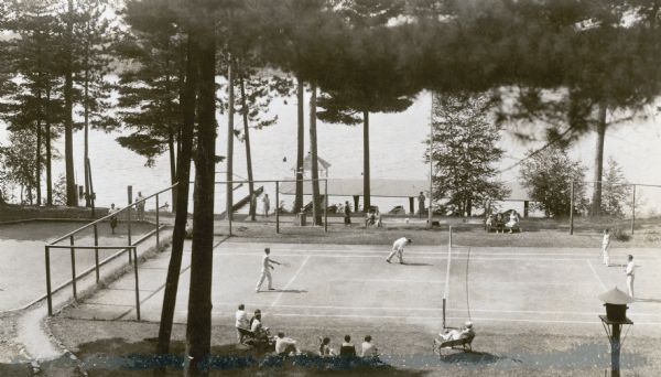 Elevated view of a tennis and a shuffleboard court, with Plum Lake in the background. Four men are playing doubles tennis, while other men are playing shuffleboard on the court behind them. There are tennis spectators in the foreground. Two piers are on the lake, and one has a covered gazebo at the end of it. There is a roof like structure parallel to the shore which protects boats moored beneath it. A caption at the bottom of the print has been obscured.  