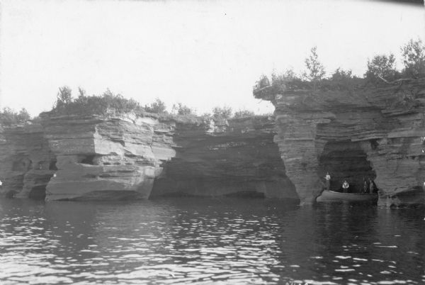 View across water towards three men and a woman posing while standing in a wooden boat in a sea cave in the Apostle Islands. Rock formations and other sea caves are on the left, and plants are growing along the top.