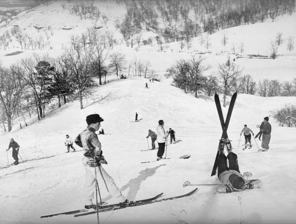 View down hill towards skiers enjoying a sunny winter day on a ski hill in Wisconsin. One man is relaxing by lying on his back with his skis crossed up in the air. There is a fence further down the hill crossing the slope from left to right, and several stands of trees.  