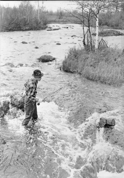 Elevated view of a man wearing a plaid shirt, waders, and a hat with flies tucked in the band casting into a stream. The caption on the negative envelope reads: "Trout fishing in the swirling white waters of a Wisconsin trout stream."