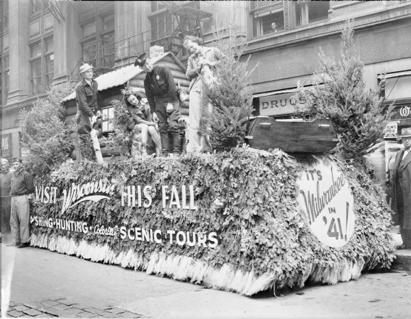 The Wisconsin float in the 1939 American Legion Convention parade features a log cabin and a boat and is decorated with small evergreen trees and boughs. There are two men and two women aboard; one of the men is dressed as a lumberjack and the other as a law enforcement officer. The woman on the right is standing and holding a very large fish, most likely a muskie. The signage on the side of the float encourages onlookers to "Visit Wisconsin This Fall. Fishing • Hunting • Colorful Scenic Tours." At the front of the float is a sign: "It's Milwaukee in 41!," a reference to the 23rd National Convention of the American Legion, which was held at the Milwaukee Auditorium in 1941. A man is looking down at the float from a second story window; there are two members of the American Legion at left and a man in a suit at right.