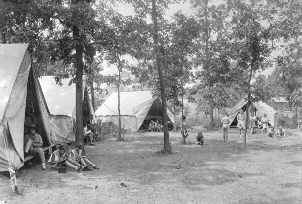 Four large canvas tents form a semicircle in an open wooded area near a lake. Girls Scouts, some in uniforms and other in casual summer attire, are posing to demonstrate camp activities such as writing or drawing, photography, and campfire starting. There is a box camera on the ground near the girls in the foreground on the left. There is a permanent structure in the far background on the right.