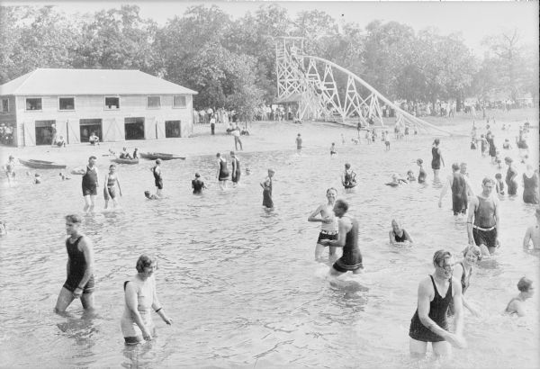 Men, women and children in modest swimming suits enjoy splashing and playing in the water at Sandy Beach, a resort established by Gerhard Strauss in 1921. There is two-story boathouse on the shore in the background, and on the right is a large wooden slide, known as the water toboggan. There are many onlookers and parked cars in the far background under trees. The resort was sold to Otto Metzker in 1934.    