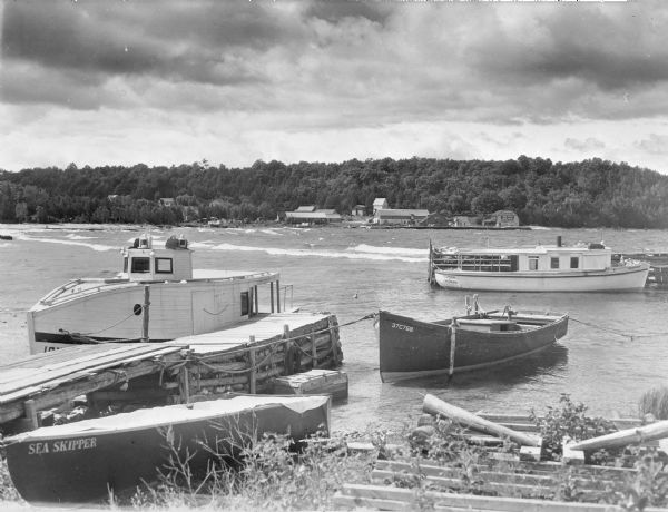 Three boats, including the <i>Sea Skipper</i>, are tied to a wooden pier near the beach at Sister Bay. Another craft, the <i>Flussheim of Sister Bay</i>, is tied to a pier beyond the others. In the background on the opposite shoreline are the buildings of the Roeser lumberyard and mill, and houses on the hill among the trees above. 