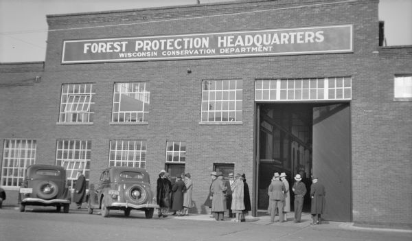 View across street towards men and women standing in small groups outside the brick Forest Protection Headquarters building of the Wisconsin Conservation Department. There are automobiles parked near the entrance. The group, members of the Association of State Foresters, have gathered for a tour.