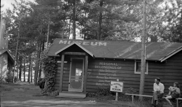 A small cottage with a tarpaper roof houses the "Gangster Museum" on the grounds of Little Bohemia. The corner of the main lodge is at far left, and Little Star Lake is in the background. A man and girl are sitting on a bench in front of the cottage. A sign painted on the siding advertises the "Personal Belongings of John Dillinger, Tommy Carroll, Homer Van Meter, John Hamilton and Baby Face Nelson." The museum commemorated an ill-fated F.B.I. raid at Little Bohemia on April 20, 1934, in which an agent and innocent bystander were killed, and the gangsters escaped unharmed.  