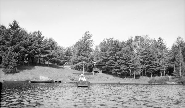 View across water towards an unidentified man fishing from a rowboat near the shore. He is wearing a light shirt and vest, and a hat. Other boats are moored along the shore. There are two cabins in the background, partially obscured by trees.
