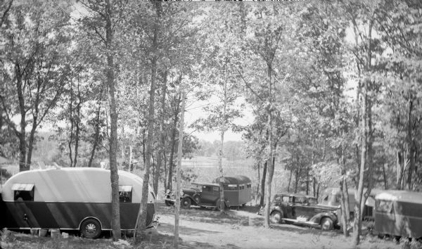 Camping trailers and automobiles are parked in a wooded area. Several trailers have awnings over the windows. The caption on the negative envelope reads" "A nest of trailer homes in the woods, Riverside Trailer Camp on the Manitowish River."