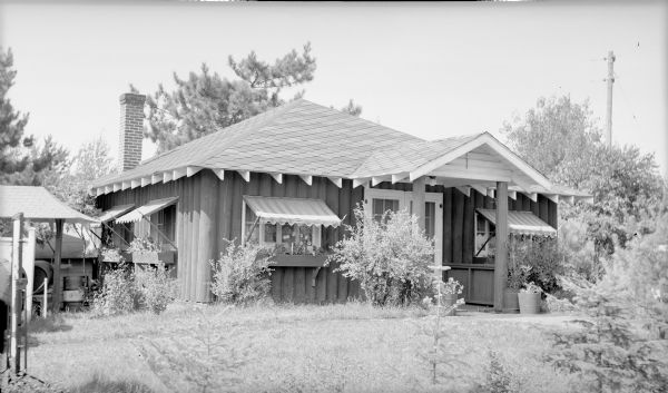 A small vertical log cabin is adorned with canvas awnings over window boxes and patterned shingle roof. There is a chimney at the rear, and the front double doors are protected by a gabled overhang. The typed description on the negative envelope identifies the building as the "Tower Tea Room Cottage, Dam Lake."