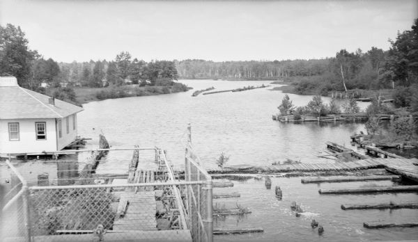 Elevated view of the fish hatchery building at Manitowish Waters, seen on the left, with a portion of the rooftop sign. The log dam and "fish way" are in the foreground, and weirs are seen in the distance.  