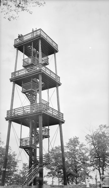 View looking up towards people enjoying the view from the top of the wooden observation tower at Potawatomi State Park. Two women are standing in the parking lot near an automobile on the right. The 75 foot tower was built in 1932.