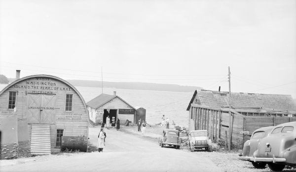 Men and women are standing around the ferry landing at Gills Rock. Automobiles are parked along the drive. Fishing nets on dryers are stacked at left, in front of a wooden Quonset building. Painted on the building are signs which read: "Visit Washington Island the Pearl of Lake Michigan," and "Modern Hotels Good Roads Beautiful Scenery." A smaller sign states: "Don't Throw Cigarettes Around." There are two smaller buildings at right.  
