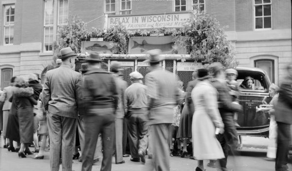 View towards a group of people looking at a float, built on the bed of a truck, representing the Wisconsin Conservation Department. The float is stopped on a street in Greenwich Village. A sign at the top of the float reads: "Relax in Wisconsin... Where Friends and Nature Meet," along with two large mounted fish, a walleyed pike and a muskellunge. Screens and signage on the side of the float draw attention to live animals, including wolves and foxes held within. There is a woman in the passenger seat of the truck who appears to be talking to a young man wearing cap and a long apron who is carrying a bugle.