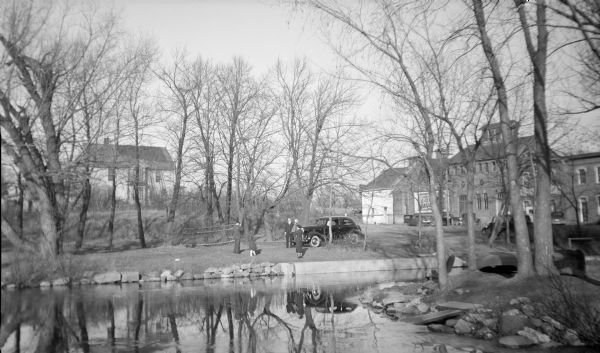 View across water towards a well-dressed man and woman posing in front of a car while another man takes their photograph. A woman and young girl are looking on. The scene is reflected in the Yahara River. In the background, right, is the Hausmann Brewing Company malt house. There is a large house in the background on the left.  
