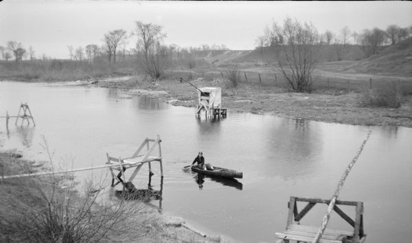 Elevated view from shoreline towards a boy using a single paddle to propel a small rowboat on the Sheboygan River. There are five rigs set up along the shores to net smelt. There is a fence and hills along the opposite shoreline.
