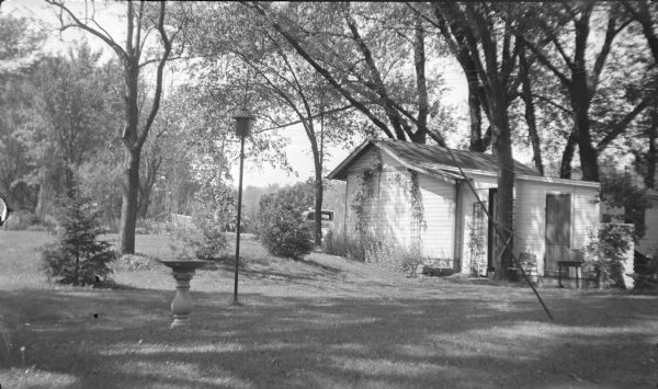 Vines are growing on trellises on the side of a small cottage on the Rock River. There is a birdbath in the yard, as well as a birdhouse on a pole. An automobile is parked in front of the cottage. The description on the negative envelope reads: "Attractive back yard of a summer home along the Rock River just north of Janesville."