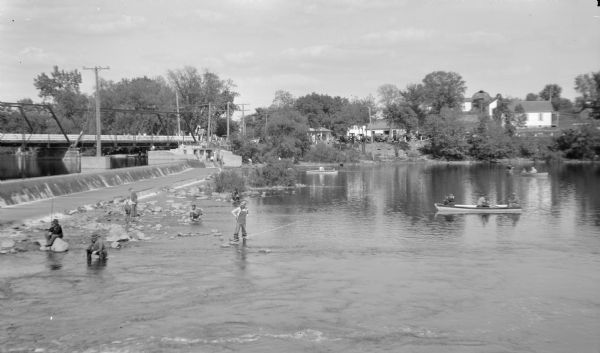View across water on a sunny day, where many anglers are fishing below the Indian Ford dam and bridge on the Rock river. Some of the men are fishing from the exposed rocks near the dam, and some men are in canoes and rowboats downstream. A Sinclair service station is on the far shoreline, and automobiles are parked near Drew's Resort.