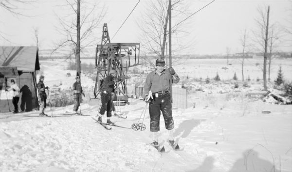 An unidentified skier wearing gaiters is posing holding his poles in his right hand and the ski tow bar with his left. Other people are lined up behind him, waiting their turn. One of the support towers for the tow is in the background. At left is the temporary wood frame shelter house which was later replaced.  