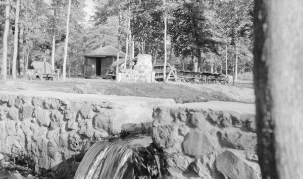 View across stone dam, with water flowing through the notch at Ojibwa Roadside Park. An automobile is parked near a large stone outdoor fireplace. There is a small cabin in the background. Rustic benches and picnic tables complete the scene.