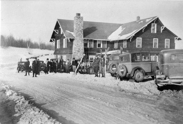 View across snowy road towards men and women posing in front of Idlewild Lodge, a two-story wood framed structure with log siding on the first floor, and a large fieldstone chimney. Three of the men are holding pairs of skis, and other skis are leaning against parked cars. Two of the cars have snow chains on the tires. A description on the negative envelope describes a "Bunch of [the] Rhinelander gang and kids out for Sunday dinner at Young's." An added notation explains, "Idlewild Lodge, Harshaw, Oneida County, Wis. (identified 1987 by an employee of the resort, which is still in operation,  Kathy Verdi.)" Edward C. Young built the resort in the early 1920's.   