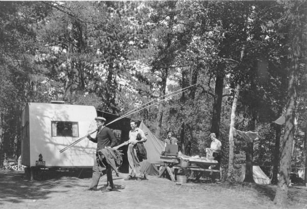 Two smiling men, one carrying long fishing poles and the other a set of paddles, are walking away from their campsites while two women are working at a picnic table. One of the women is wearing a stylish hat and is standing behind a camp stove, and the other woman is drying dishes. A camping trailer with an Illinois license plate is parked on the left, with large awning extending from its side. There is a tent in the background on the right. Clothes are drying on a line between two trees.