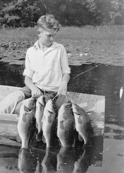 View across water towards a boy sitting in the stern of a boat holding a stringer of five good sized fish, their tails in the water. A lure is hanging from his fishing rod. There is a bed of waterlilies along the shoreline in the background. The caption on the negative envelope states: "Indeed proud of his day's catch of black bass!"