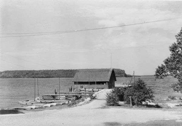 Canoes and small sailboats are pulled up on the shore or tied up to the piers extending from the wharf at Ellison Bay. There is a small warehouse on the wharf. Ellison Bluff is along the shoreline in the background.