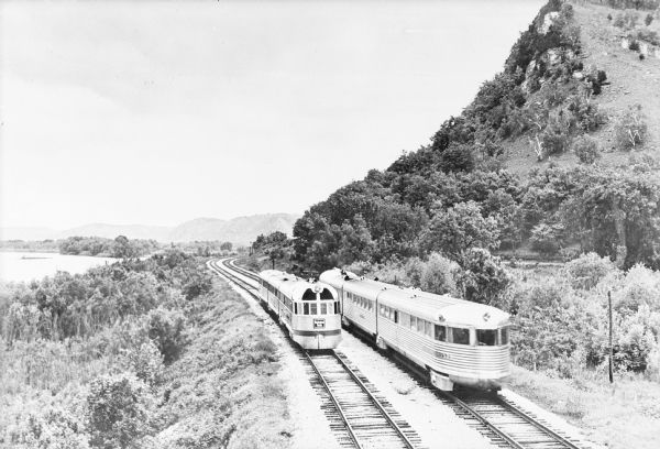 Two streamlined style, stainless steel clad passenger trains meet on the tracks at the base of a bluff along the Mississippi River near La Crosse.  A sign on the front of the oncoming train reads: "Burlington Route."