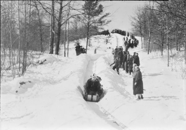A young man is steering a toboggan with three others on board down a long course.  Spectators line the run on the right.  