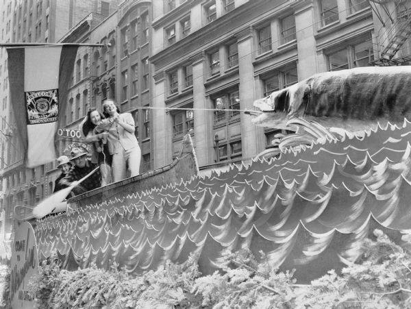 A Wisconsin Conservation Department float in the 1939 Chicago American Legion Parade. The float features two women who are standing in a canoe pretending to pull in a muskie with a fishing pole, which is attached by a fishing line to a large sculpture of a muskie at the front of the float. Two men, one with a paddle, are sitting in the back of the canoe. The sides of the float have been cut and painted to resemble water, and evergreen boughs simulate a shoreline. A sign on the side of the float reads: "Come to Wisconsin and Catch a Muskie." A banner hanging on a pole in the left foreground reads: "Welcome, Legionnaires."