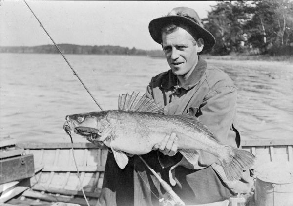 An unidentified man, wearing a hat with fishing flies on the hatband, is posing in a canoe holding a walleyed pike. Several hooks are protruding from the fish's mouth. A caption on the negative envelope reads simply: "A large pike caught in a southern Wis. lake."